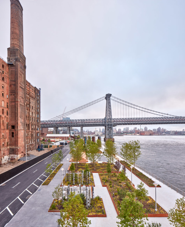 The Domino Sugar Refinery rises behind the park. The park's central area features planters with shade and flowering trees interspersed with Hanover concrete slabs that separate into pathways.