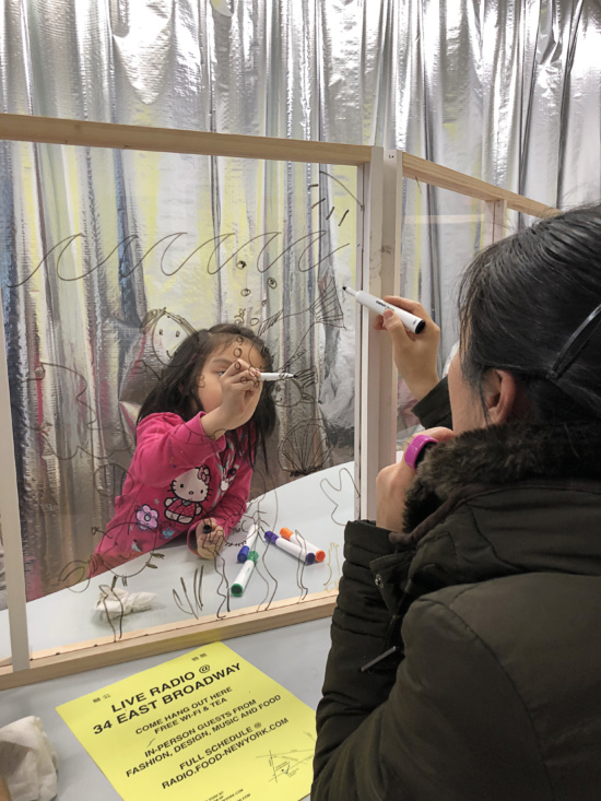 A child and an an adult draw with dry-erase markers on opposite sides of a glass pane.