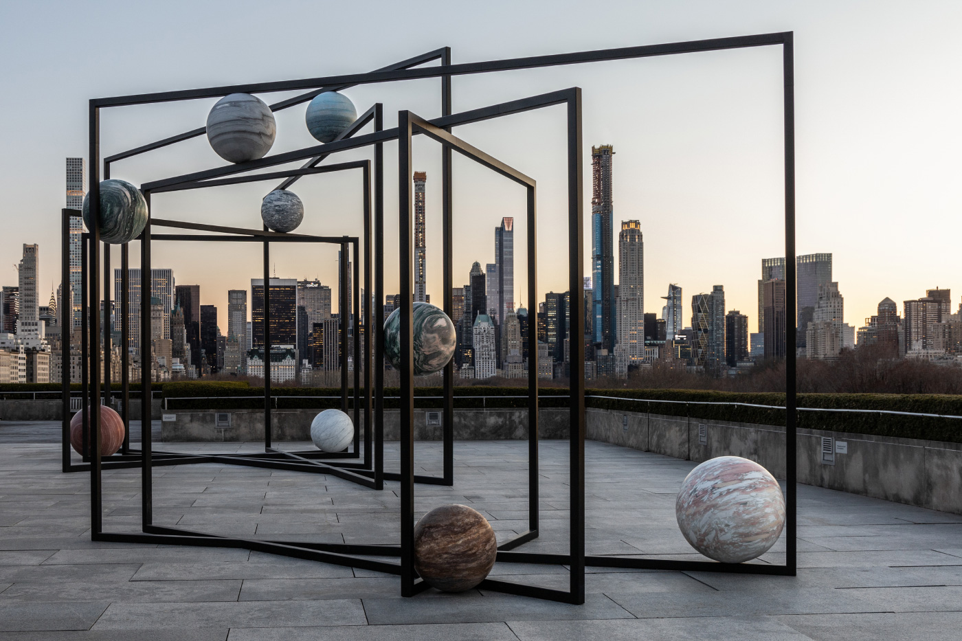 Photo of large stone spheres of different colors, suspended in black steel frames, against the Manhattan skyline