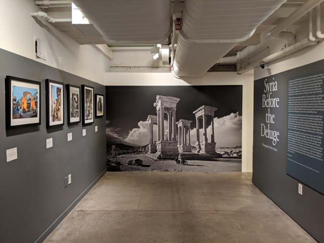 Photo of a small gallery interior with a photo on the wall and "Syria Before the Deluge" written on the wall