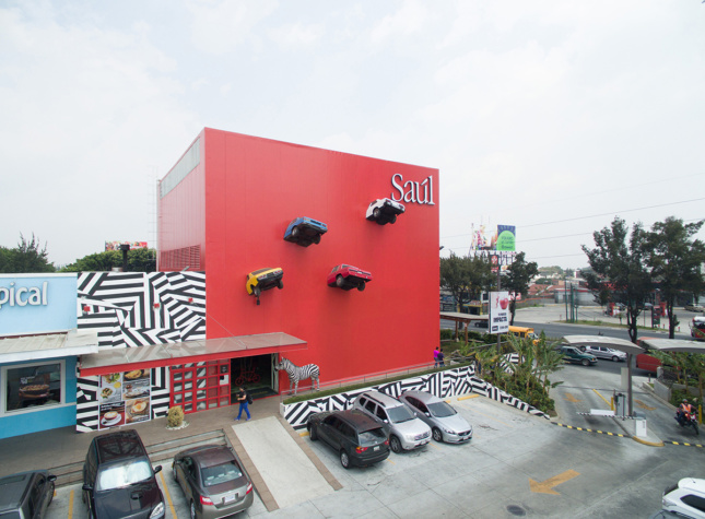 Photo of a large red, box-shaped building with the fronts of cars attached to it