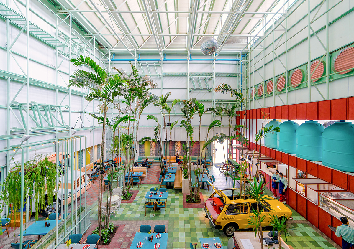 Photo of a room with a very high ceiling with bright colors, palm trees, and restaurant seating inside