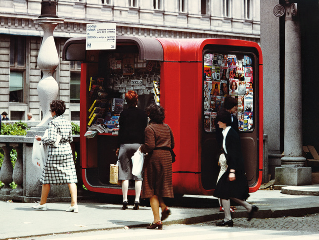 Photograph of a rounded red kiosk selling magazines on the street with people walking by it