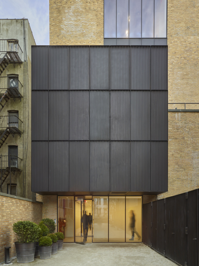 Photo of a blank gray facade hovering over a ground-level glass-walled entrance