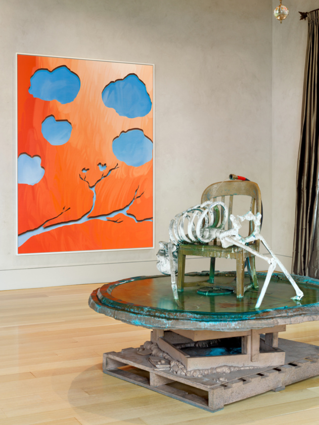 A sculpture of wooden palettes topped with a fountain props up a skeleton on a chair, with a garden hose on top. In the background a painting of a tree black and clouds—the sky is orange and the clouds and branch are produced in negative in blue.