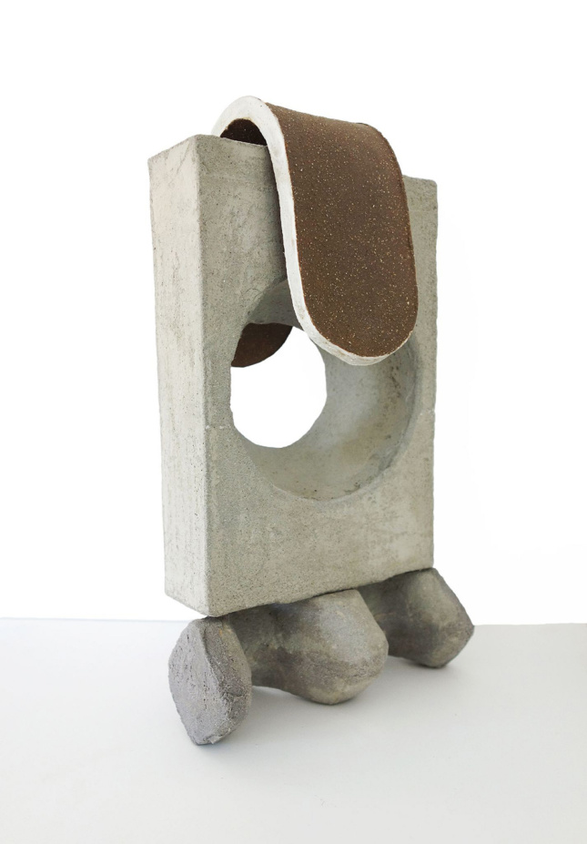 A close up photo of a sculpture—a piece of rectangular concrete with a circular hole mounted on top of a tripartite concrete form with a thick, brown glazed ceramic tongue-like ovoid draped over the top.