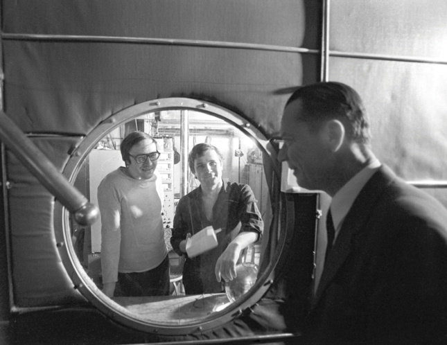 Black and white photo of two people looking through a porthole at an observer