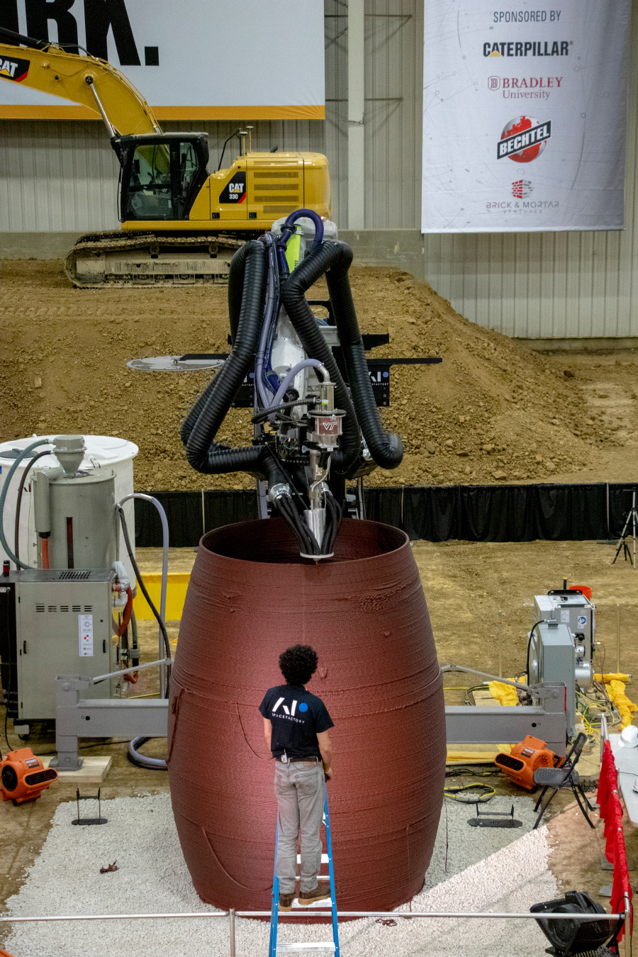 Photo of a person standing in front of a cylinder that's being 3D printed, a model for space habitation
