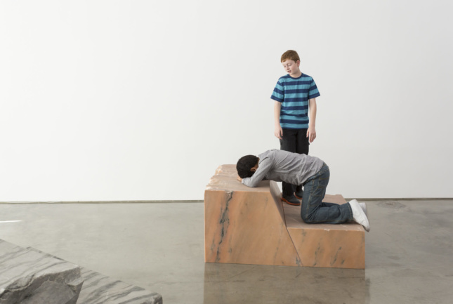 Photo of art installation with one young boy kneeling on a marble pedestal with another boy overlooking him