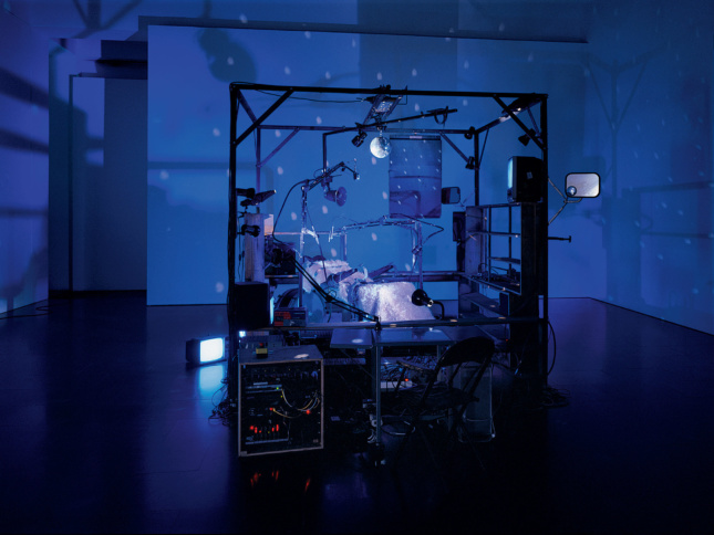 Photo of a gallery lit with blue light and an installation of a bed with various devices around it