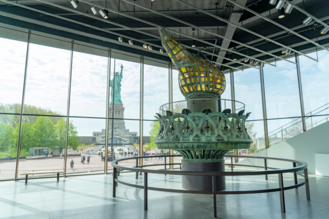 Photo of a multistory copper torch foregrounded against the Statue of Liberty