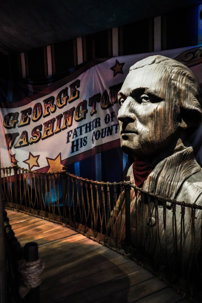 A large wooden bust of George Washington sits next to a banner that reads "George Washington, Father of his country"