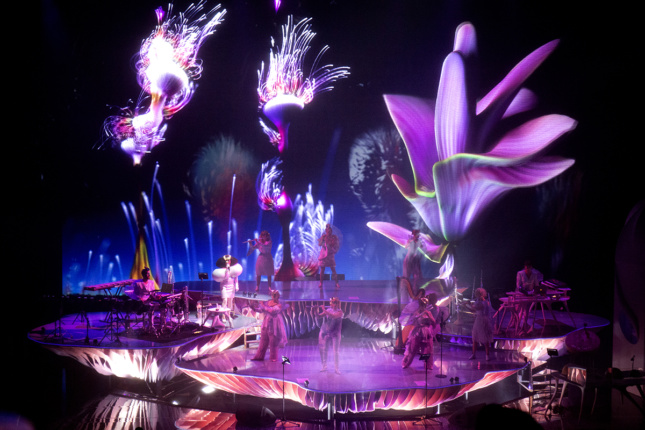 Musicians perform on purple platforms while imagery of fireworks and flowers are project large behind them.