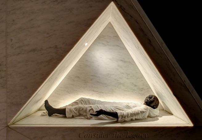 Photo of a sculpture of a black man reclined in a triangular cut-out structure.