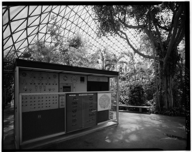 Black and white photo of the interior of a geodesic dome with a hut and many plants inside
