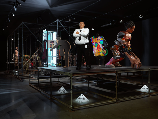 Photo of figurative sculptures of a man in a suit, a giant automaton, and others are on a large platform.