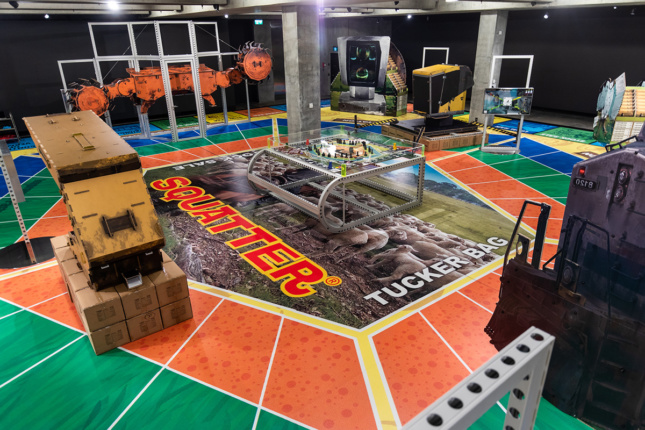 Photo of a large board game with a central image over some sheep and text reading SQUATTER has a variety of life-size realistic sculptures of mining machines on it.
