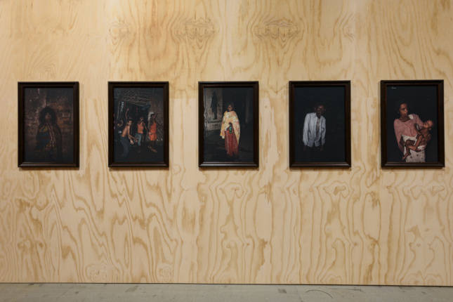 A plywood wall with five portraits on it, each depicting a disheveled homeless individual on a black background