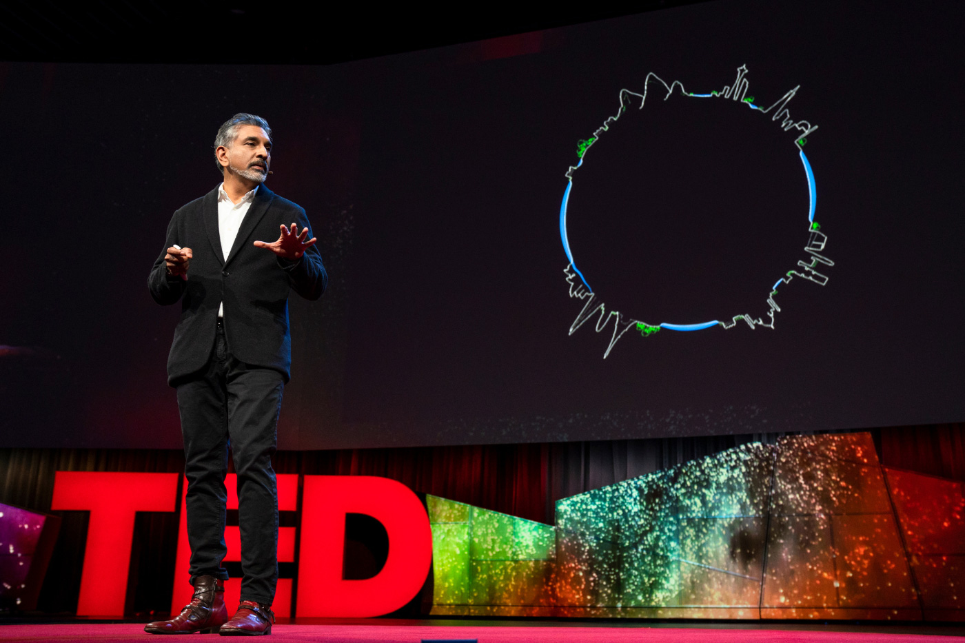 An indian man in a black suit on a stage, with the letters TED in large text