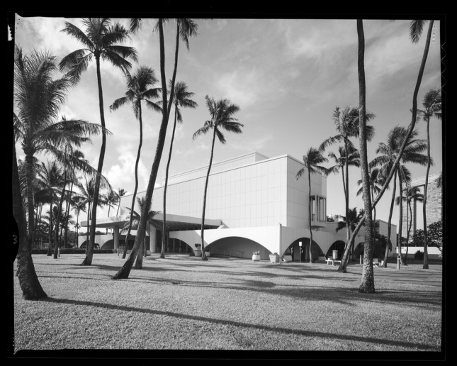 Black and white photo of a boxlike performance venue with palm trees in foreground