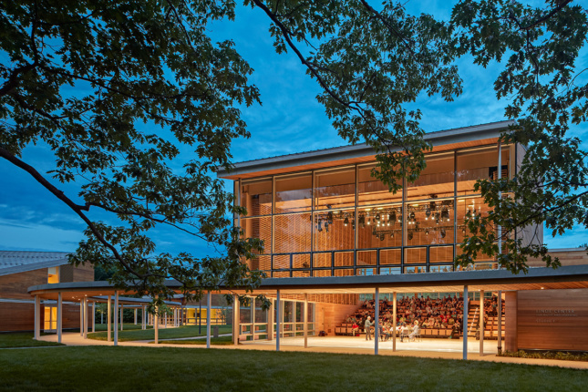 Twilight image of two story structure with performance lighting up interior, and pavilion leading to other structures