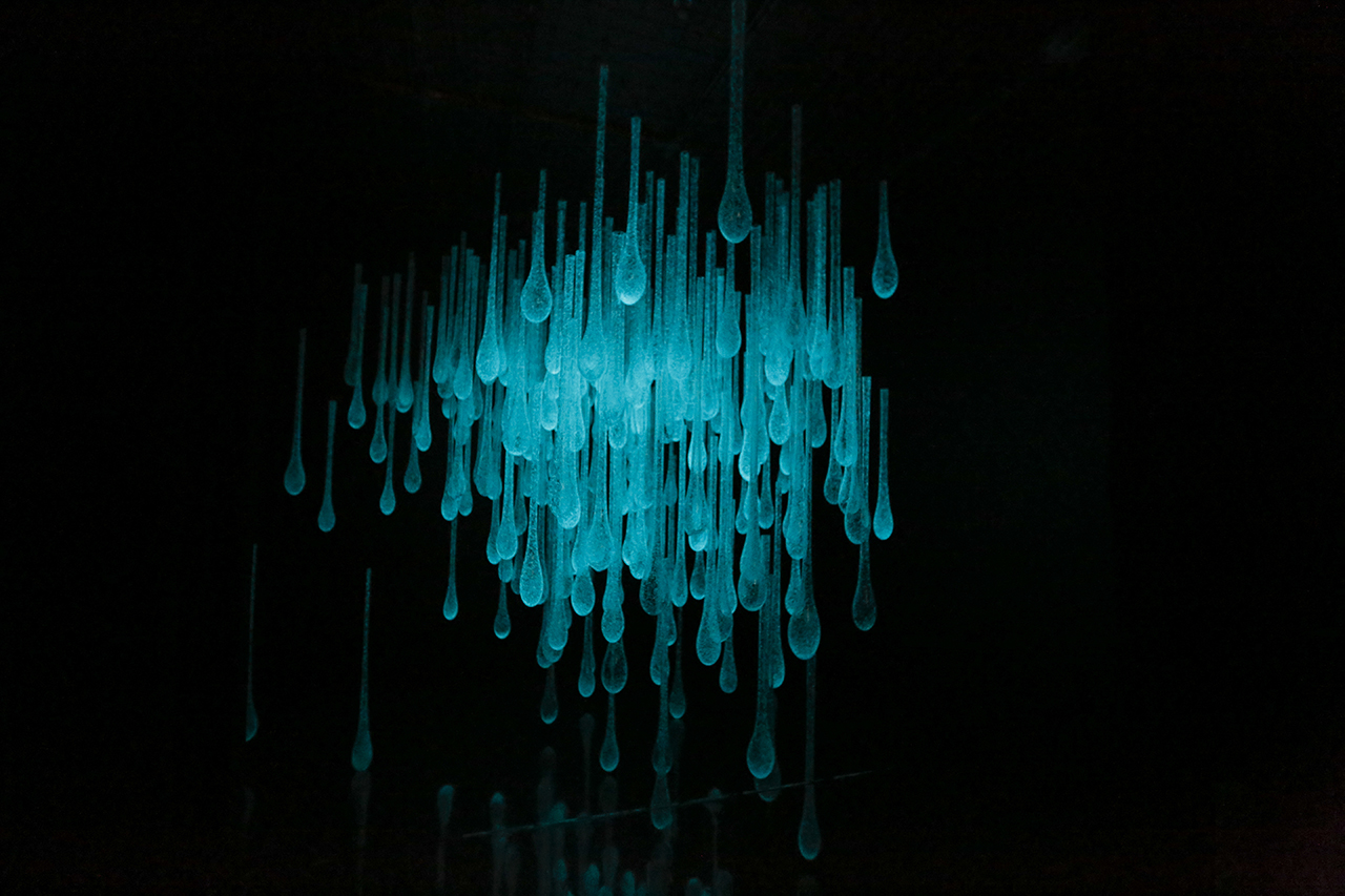 an installation in a dark space, with glass vessels hanging down from the ceiling that light up each time a visitor walks into the space