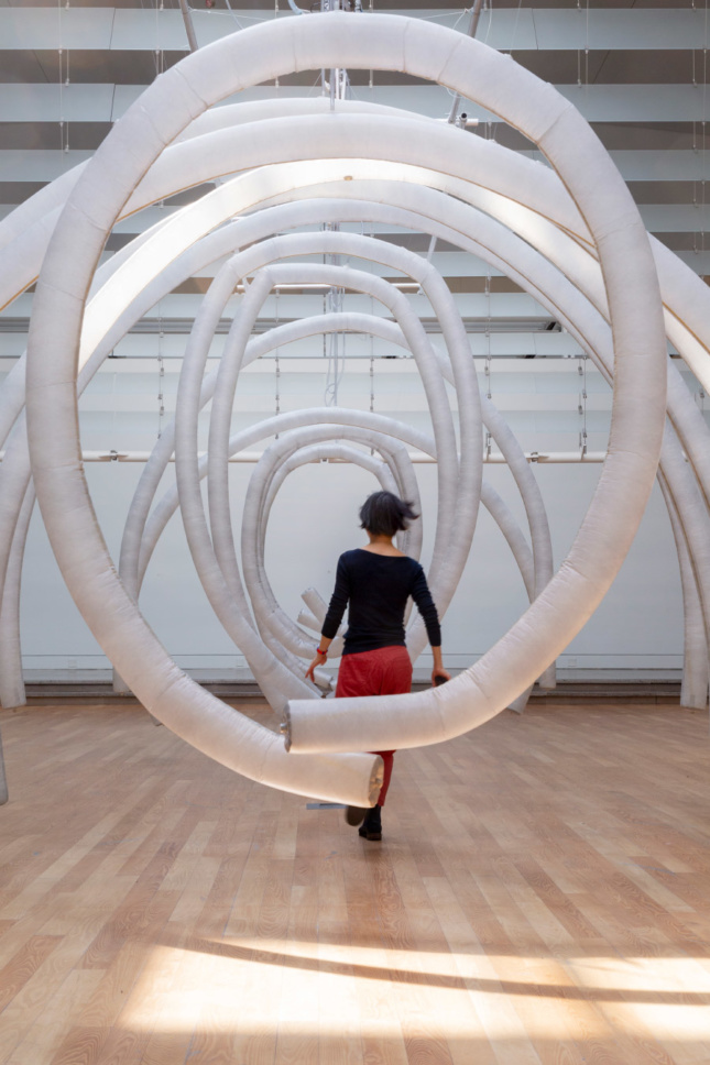 Photo of the Queens Museum atrium with inflatable ribs suspended from the ceiling, with a woman running through the tubes