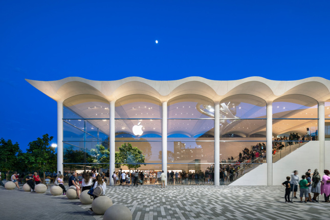 Twilight shot of glass-clad Apple store with undulating white roof situated next to mall plaza