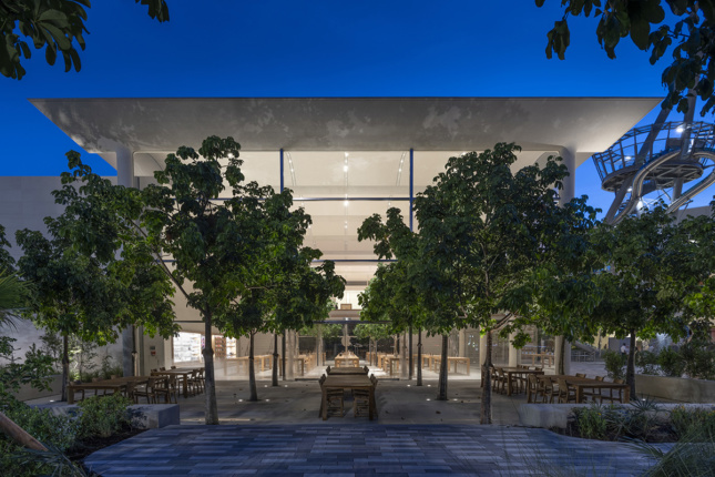 Twilight shot of courtyard with trees and wooden tables outside light-filled boxy building 