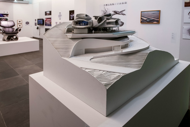A photograph of an architectural model sitting on a podium in a white-walled gallery. Photographs, drawings, and other works of art are featured on the walls and on other podiums throughout the space. 