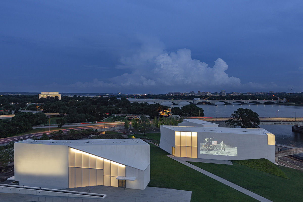 Aerial image of two angular white buildings on green campus lit at twilight by opaque glass windows, which forms The REACH