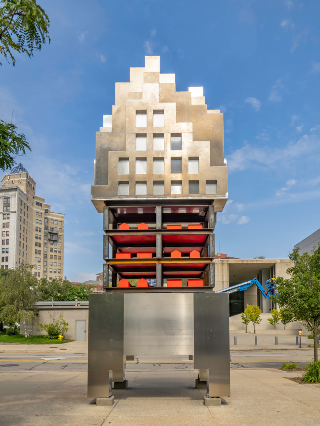 A 25-ft tall metal sculpture stands upright on a wide sidewalk. The sculpture resembles an abstracted form of a high rise building and featueres windows of various sizes. The middle of the sculpture contains a cage-like section made of metal beams and it holds multiple red house-shaped forms. 