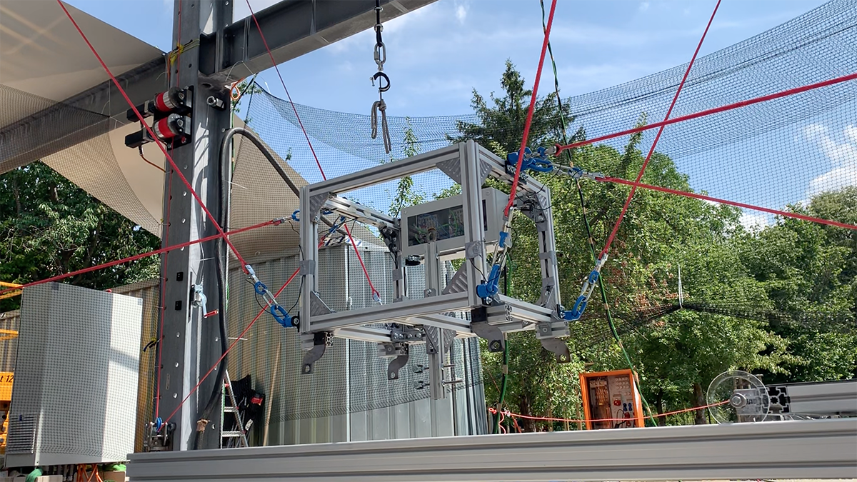 A robot in the form of a metal box is attached to various red cables and suspended a few feet in the air.