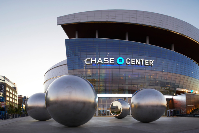 Photo of glass-clad arena building with silver spheres arranged on plaza in front 