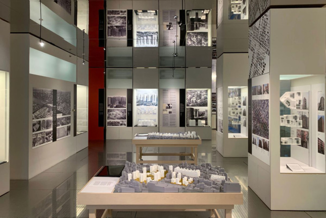 A photograph of a museum gallery filled with architectural models on a table and photographs on the wall.