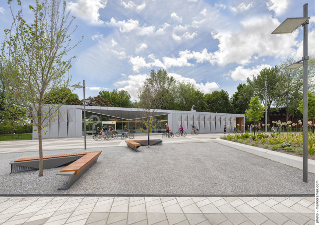 A small building with a metal facade sits in a concrete paved park surrounded by trees. 