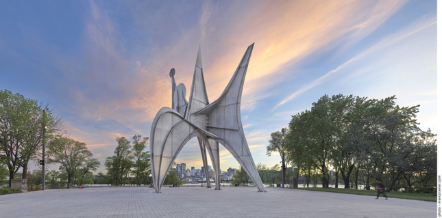 A monumental metal sculpture sits in the middle of a park with the sun setting behind the Expo 67 site