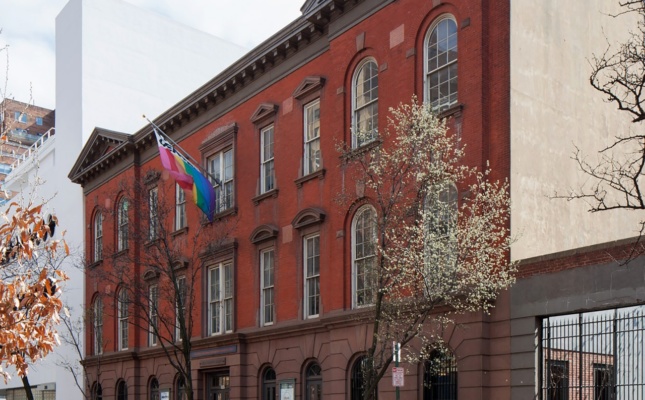 A perspective photo of a brick building on a tree lined street. A rainbow flag flies above the building's entrance.