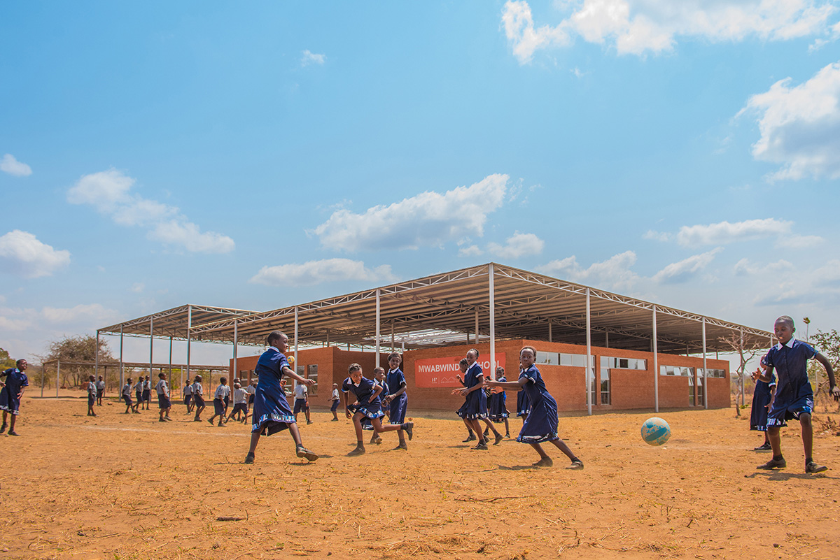 Image of African children playing soccer in front of brick school with a sign that says Mwabwindo