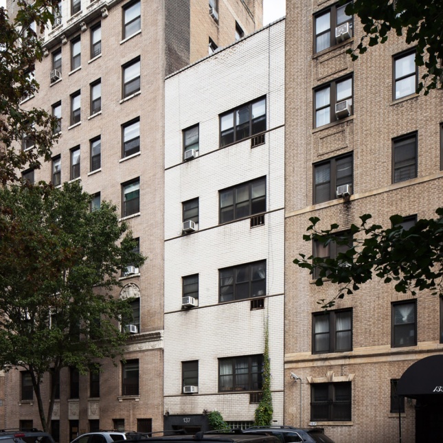 A photo of a modern, white, brick apartment building surrounded by taller apartment buildings on a tree-lined street.