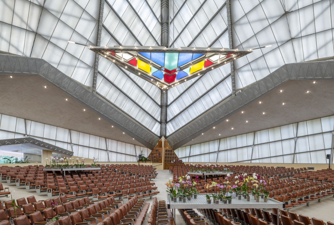 A stained glass triangle hangs under a pyramidal roof over seats and flowers.