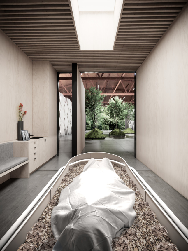 A body is shown in a pale wooden room covered in white and placed in a vessel for decomposition and covered with wood chips. A pivoting door is open in the background showing trees planted in the adjacent room. 