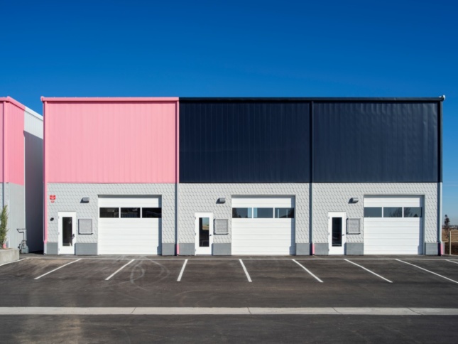 A boxy building sits in a parking lot. It has three white garage doors and covered in a grey shingle pattern with pink and black accents, designed by Paul Andersen