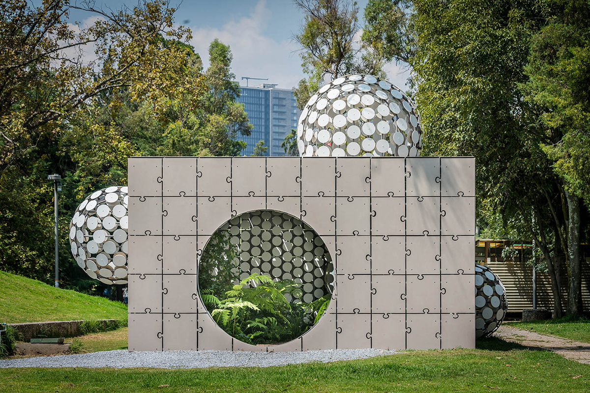 A concrete cube pavilion with puzzle piece–like panels that has bulbous forms made of white circles growing out of it.