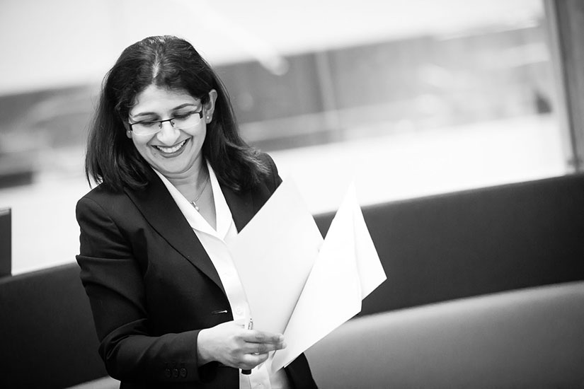 A black-and-white photo of a woman, Upali Nanda, in a dark suit smiling and holding papers.