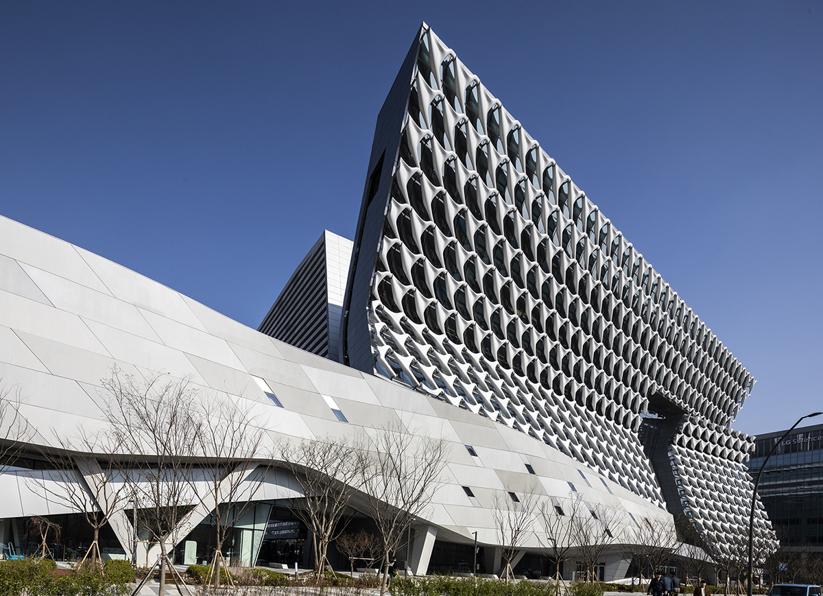 A tall building with a sweepiing facade made of interlocking white Ts, designed by Morphosis. The building continues at a lower height as a undulating gray roof with some irregular fenestration.