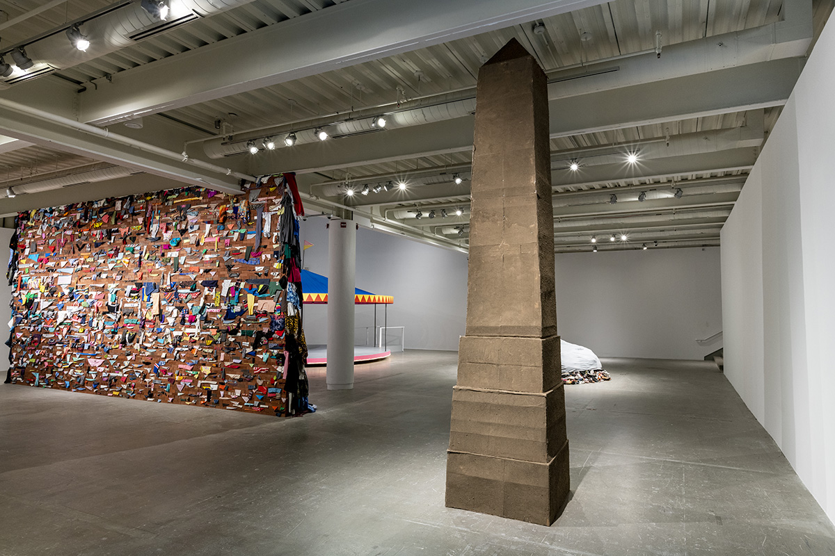 Gallery with obelisk sculpture, brick wall with wedged colorful garments, and a carousel in the background