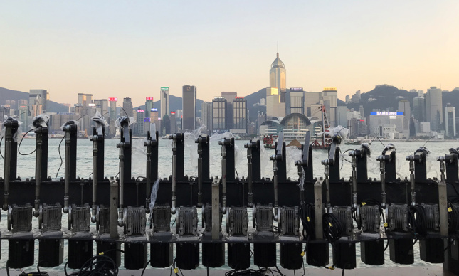 An array of robotic armatures in front of Hong Kong's harbor with the skyline in view.