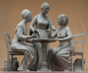 Photo of statue of two women sitting at a table and a third standing behind a table