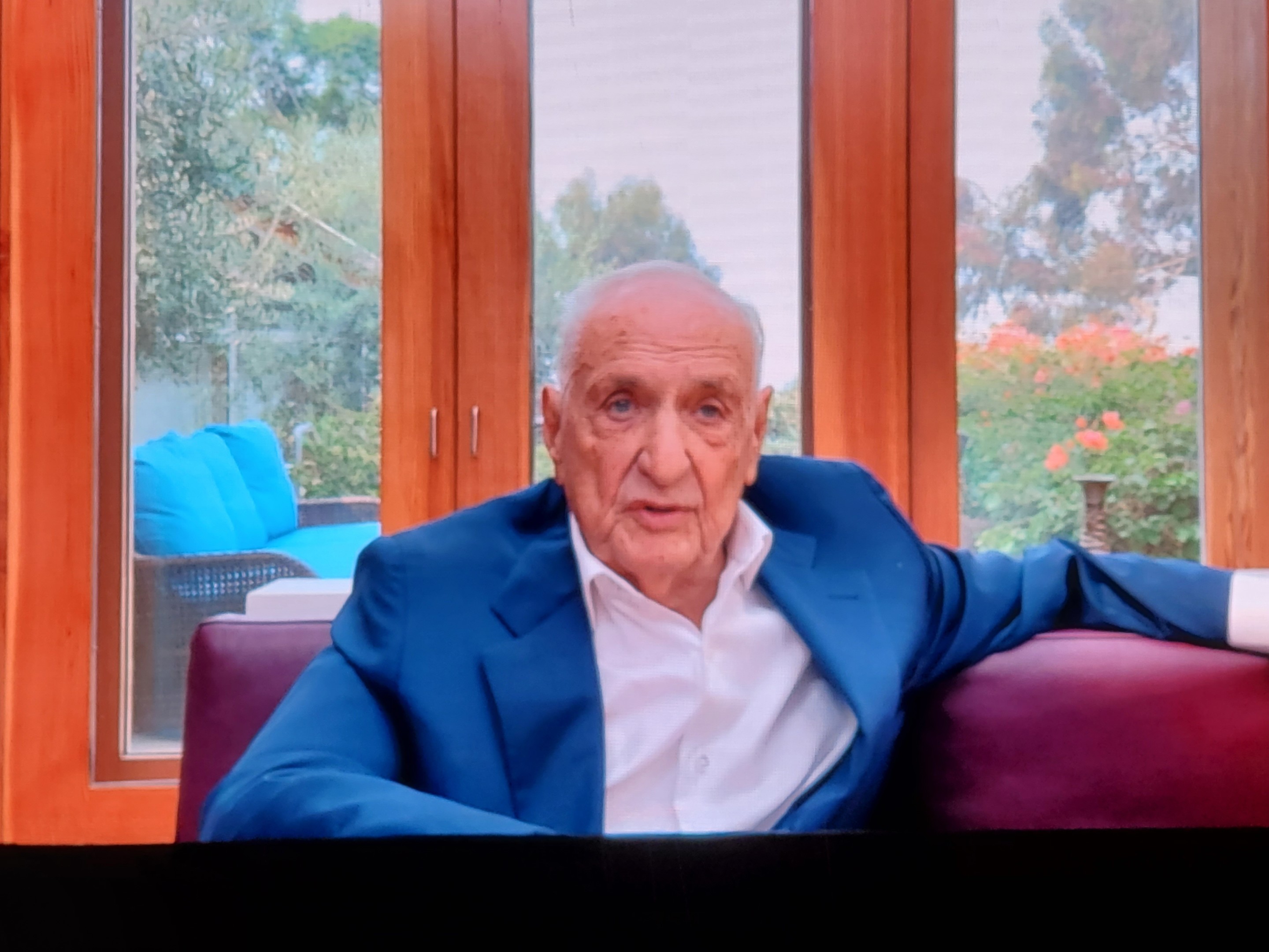 Frank Gehry, an elderly man, delivering comments on a video screen